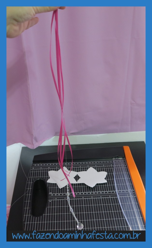 Corte 4 pedaços da fita de cetim rosa, com 20 a 30cm. Segure as fitas junto com o fio de nylon, de modo que as fitas terminem onde começa a decoração com miçanga. Ou seja, onde termina a fita de cetim começa a decoração com miçangas.